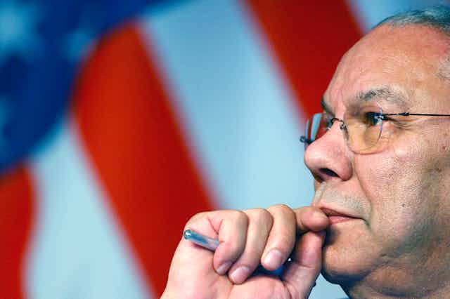 Colin Powell, a pen in hand, looks into the distance as an American flag hangs behind him. 