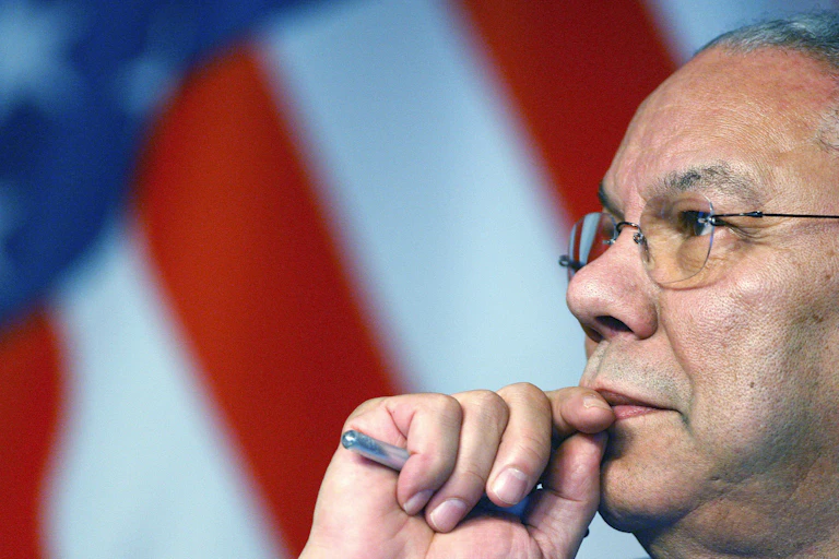 Colin Powell, a pen in hand, looks into the distance as an American flag hangs behind him.