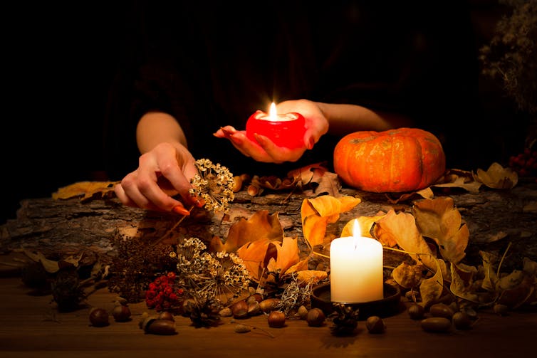 Hands with sharp red nails arrange a display of candles, herbs, pumpkin, nuts, dry leaves and berries.