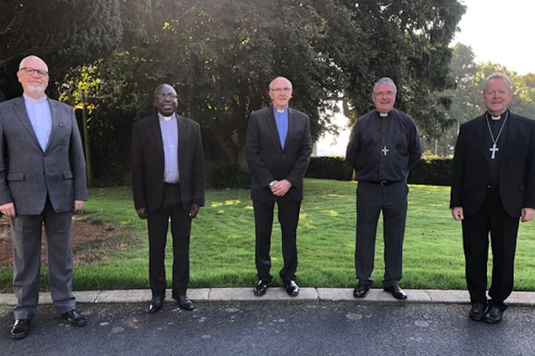 Five men wearing suits, some of them wearing clerical collars or crosses, stand in a line outside, facing the camera.
