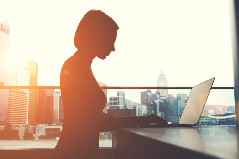 A woman is seen in silhouette looking at her laptop in front of an office window.