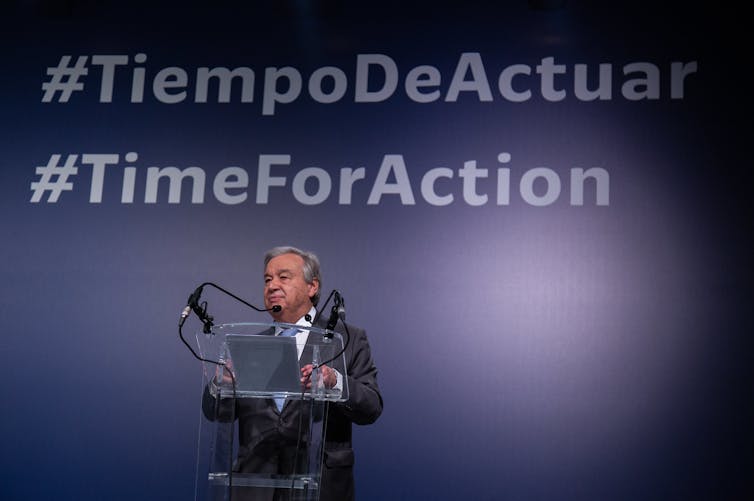 Guterres standing at a podium with #TimeForAction on the screen behind him