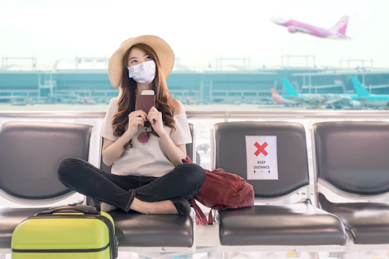 Masked female student holding passport sits in airport lounge as plane takes off behind her