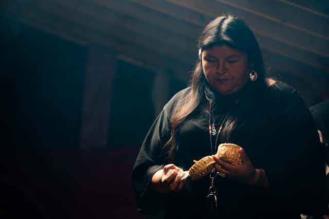 A woman holds a small cedar basket in a dark backlit room