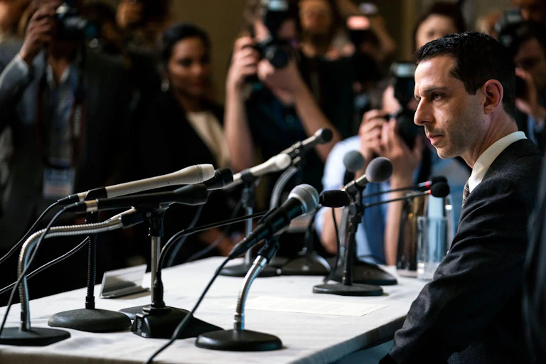man in front of microphones with press in background.