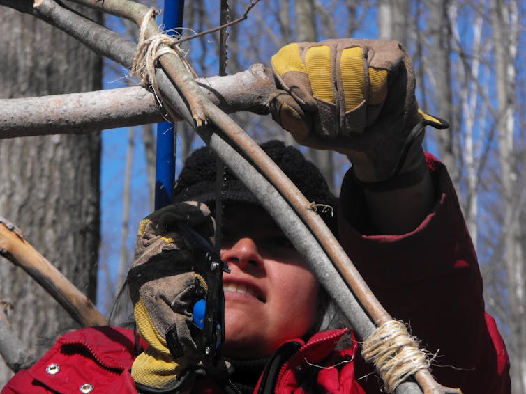 A woman holds a saw as she builds a lodge. The image is closeup, you can see her jacket (red), gloves (yellow)
