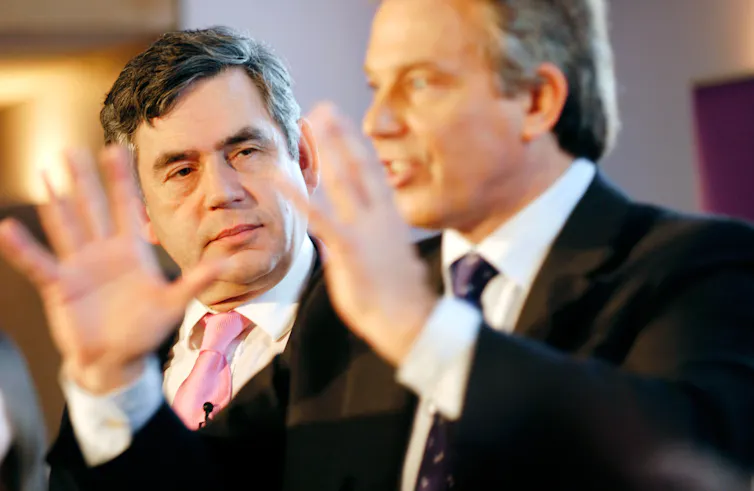 Tony Blair speaks and gestures with his hands, while Gordon Brown looks on stoically