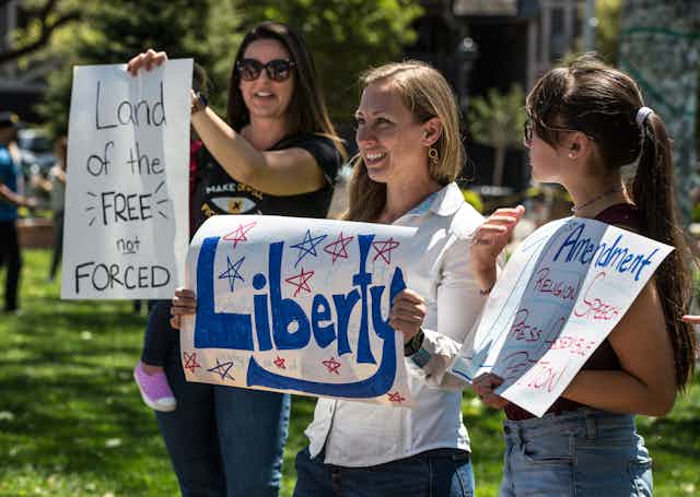 Three women holding signs that say "Land of the FREE not FORCED," "Liberty" and something about the First Amendment and freedom of religion and speech.