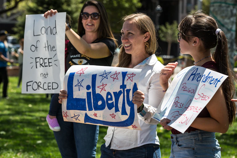 Three women holding signs that say "Land of the FREE not FORCED," "Liberty" and something about the First Amendment and freedom of religion and speech.
