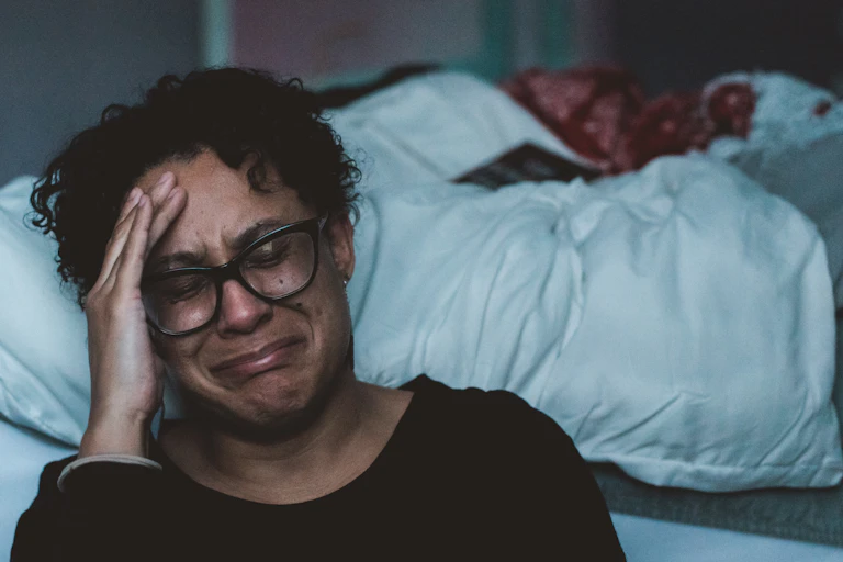 A person sits by their bed, hand on forehead, about the cry