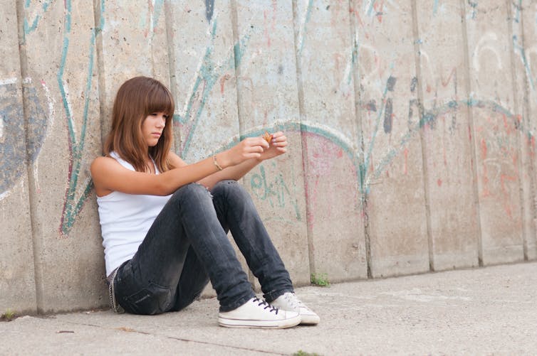 Young woman in casual clothes sitting on the ground leaning against a wall looking sad.
