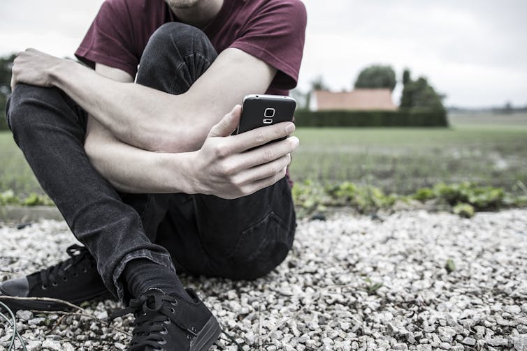 Cropped image of young person sitting cross-legged on the ground looking at a phone, with head out of shot