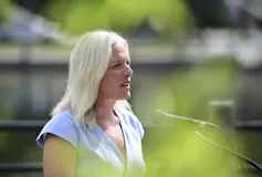 A blonde woman in a blue shirt speaks into a microphone