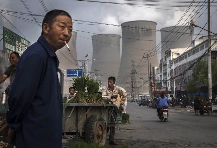 A man stands in a street market smoking, with cooling towers for a power plant behind him.