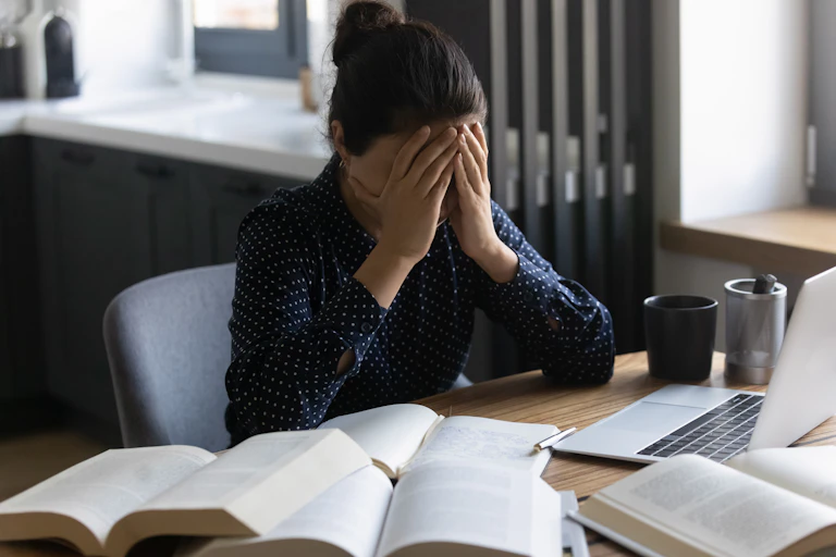 Young Indian female student with head in hands as she sits at laptop with several books open