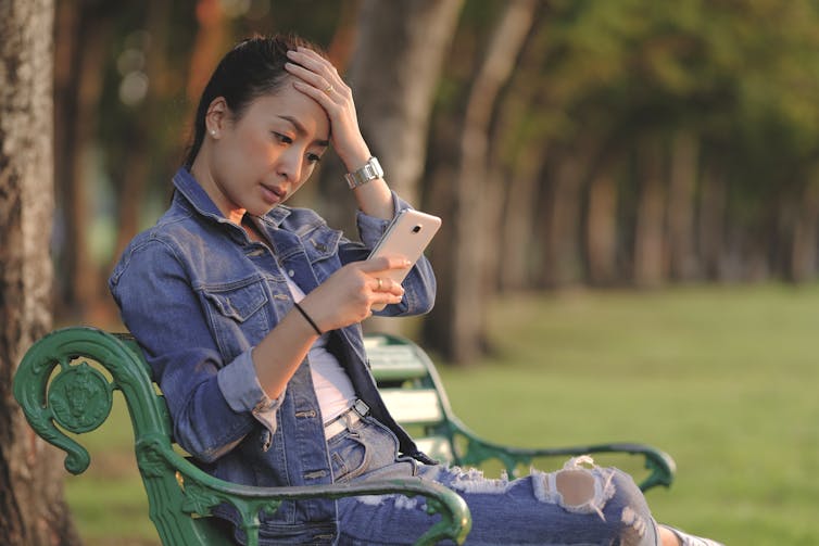 A young woman sitting on a park bench looking at her smartphone. She appears stressed.