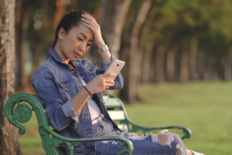 A young woman sitting on a park bench looking at her smartphone. She appears stressed.