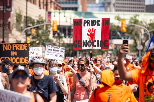 People wearing orange shirts walk holding signs that read "no pride in genocide"
