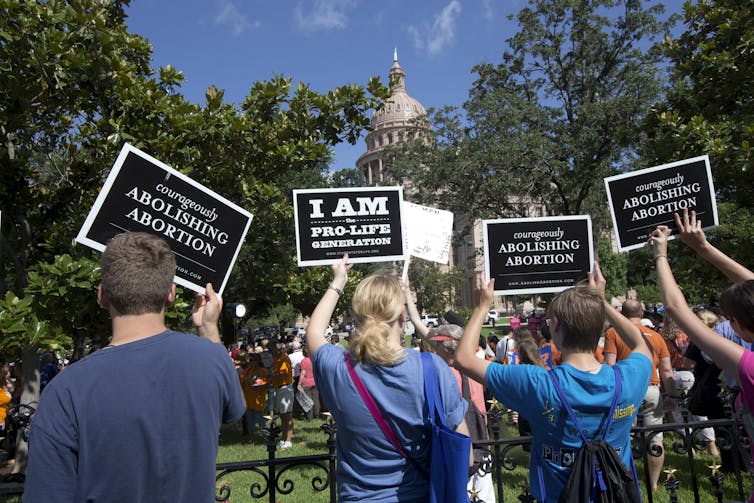 Four people face away from the camera holding up signs that read 'Courageously Abolishing Abortion'