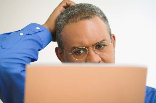 Man wearing glasses stares at a laptop screen, which obscures the lower half of his face, while scratching his head