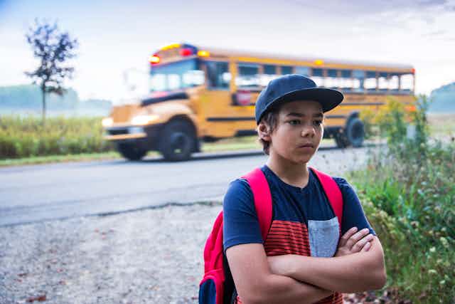 Boy wearing baseball cap and backpack stands in foreground with arms crossed while school bus drives past in background