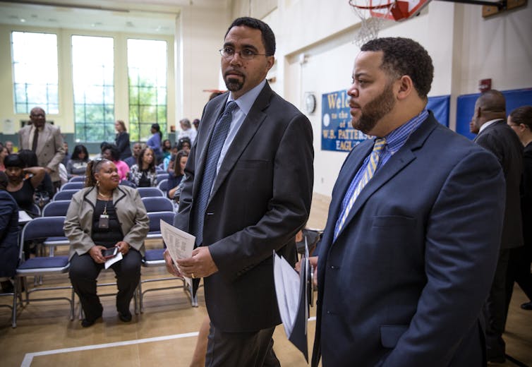 Two men in suits walk through a school gymnasium where adults are seated in rows of chairs