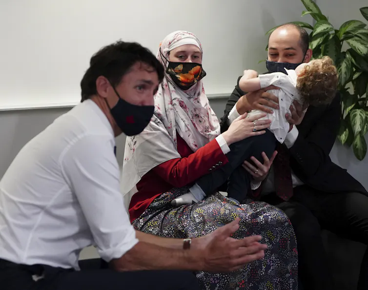 Justin Trudeau in profile wearing a face mask in the foreground, a couple with a child in the background