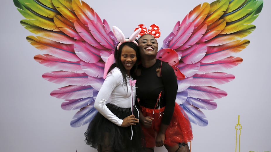 Two women smile in front of an artwork featuring oversized, colourful angel's feathers.