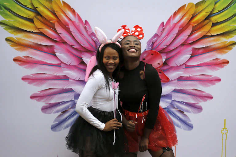 Two women smile in front of an artwork featuring oversized, colourful angel's feathers.