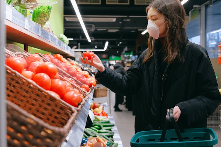 Woman in black coat and face covering holds shopping basket and looks at display of fruit