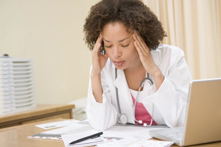 Black woman doctor wearing a white coat with a stethoscope around her neck with her fingers on her temples at her desk
