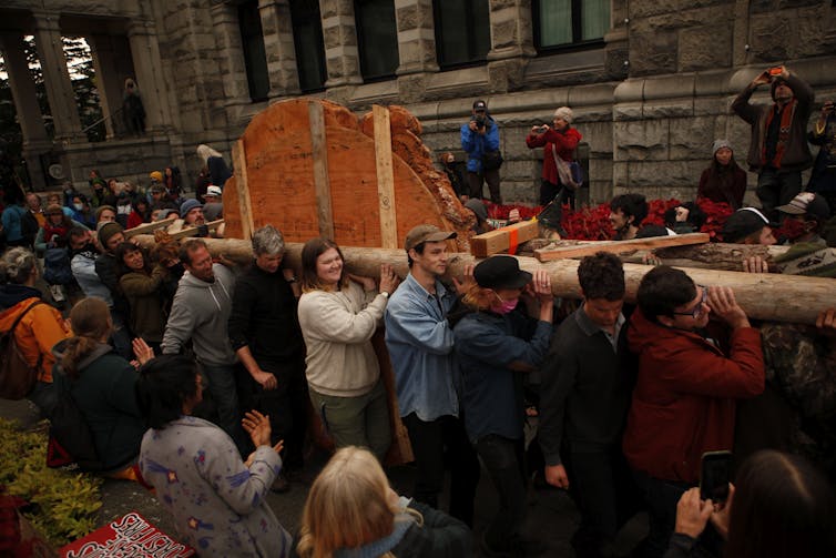 a slice of a tree is carried sideways by a group of people outside the legislature building