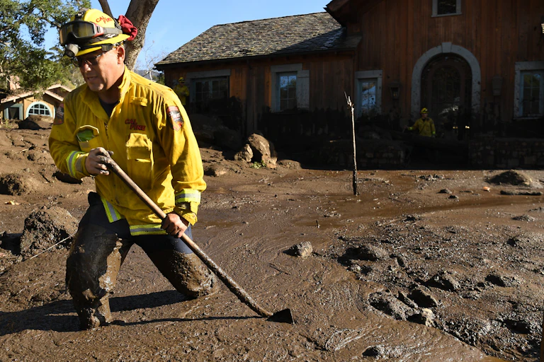A firefighter walks through mud up to his knees in the yard of a home hit by a mudslide.