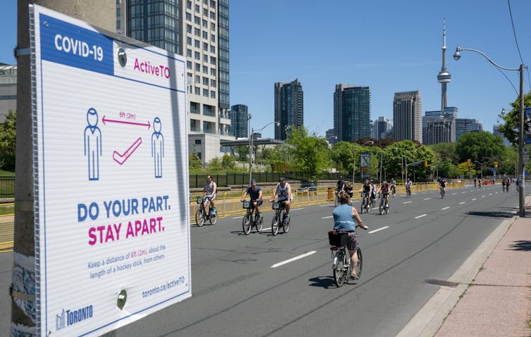 Cyclists on Lakeshore Blvd. with the downtown skyline in the background and a physical distancing sign in the foreground.