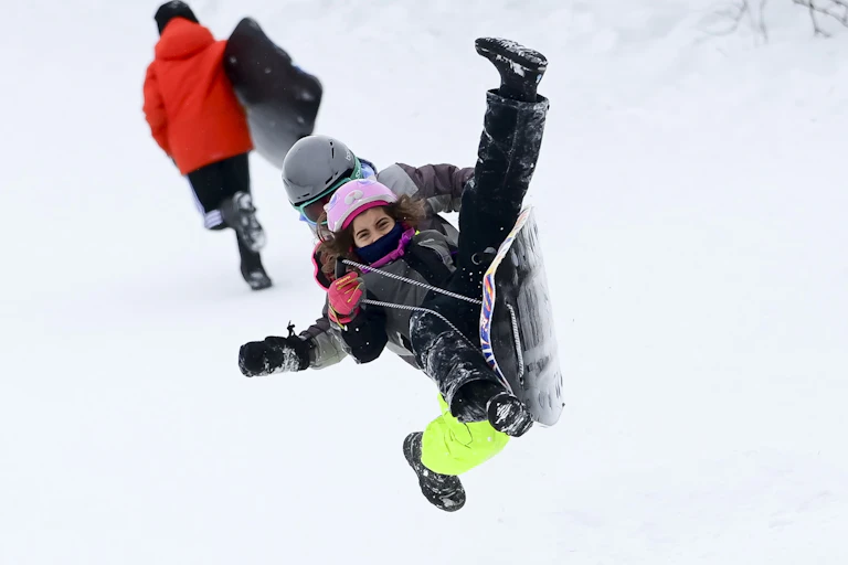 Two kids on an airborne toboggan in the foreground with an other kid carrying his sled up the hill in the background