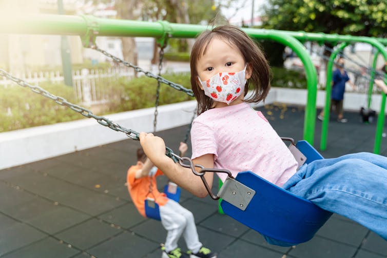 Girl in a face mask on a swing in the foreground with a boy on a swing in the background