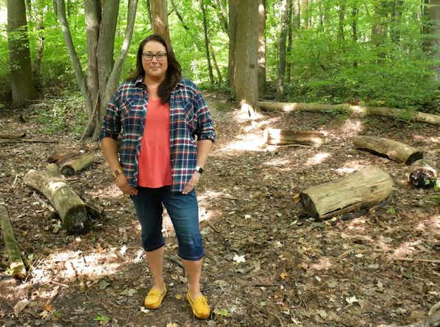 A woman stands in the forest wearing a plaid shirt, coral top and jean capris.