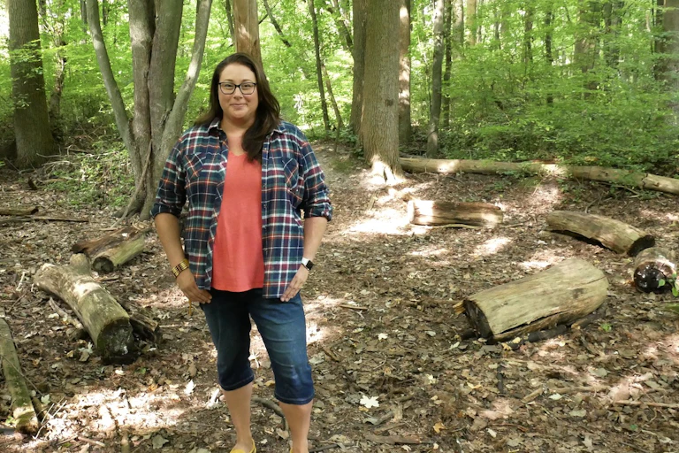A woman stands in the forest wearing a plaid shirt, coral top and jean capris.