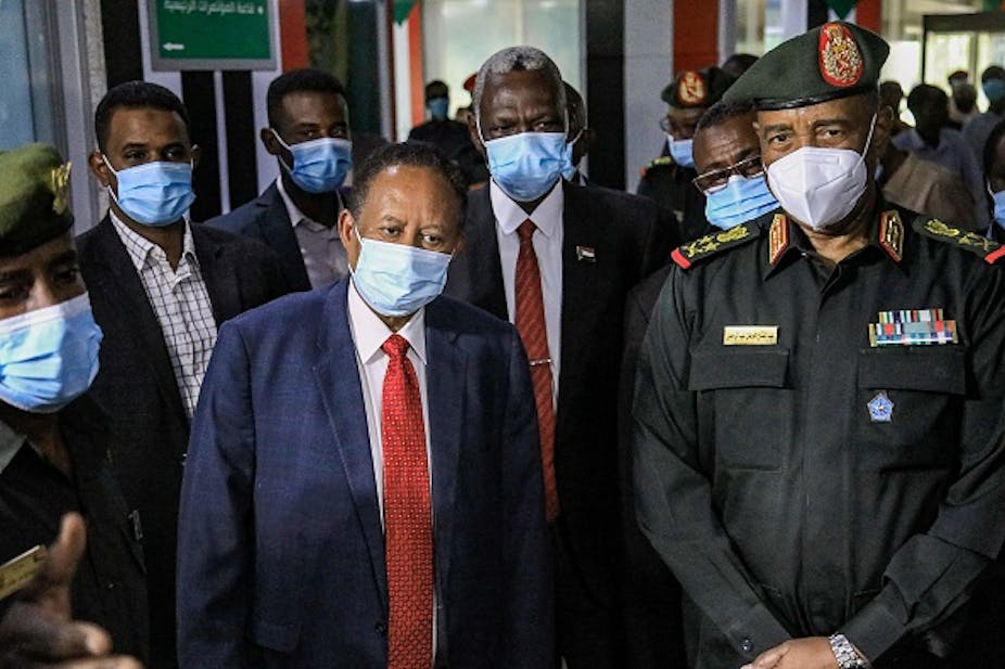 A man in a suit and face mask stands next to a man in military uniform and face mask; other men in face masks stand behind them