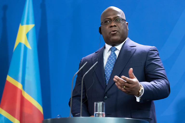 A bald-headed, bespectacled man wearing a suit and tie speaks into two tiny microphones while standing at a podium in front of a flag
