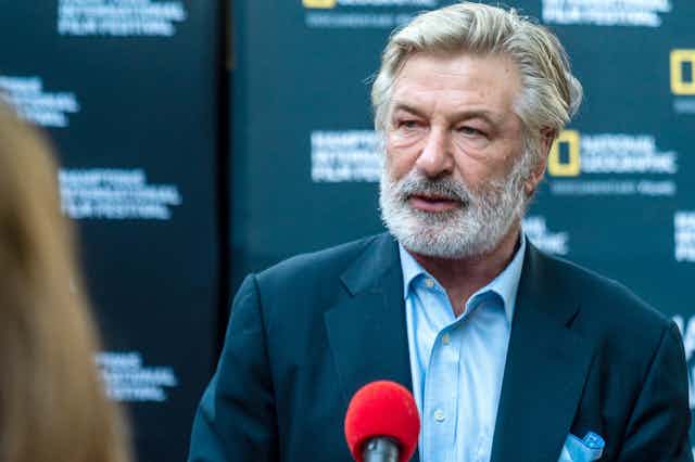 Actor Alec Baldwin wearing a suit and with a grey beard stands in front of a microphone.