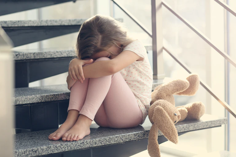 A sad little girl hides her head, sitting on the stairs, a doggie by her side