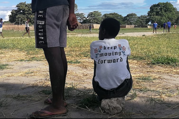 A young girl sitting on a stone in a patchy football pitch.