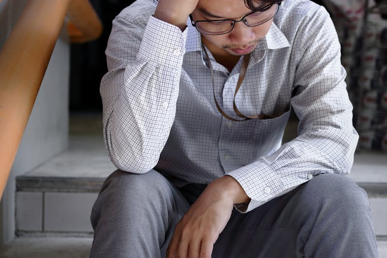 An asian man sits on the stairs with his head in his hand looking sad. He is wearing grey pants, a light blue shirt and glasses.