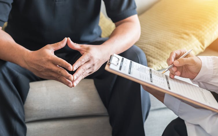 A man is seen with his arms on his knees, his hands are clasped. A doctors clipboard is visible to the right.