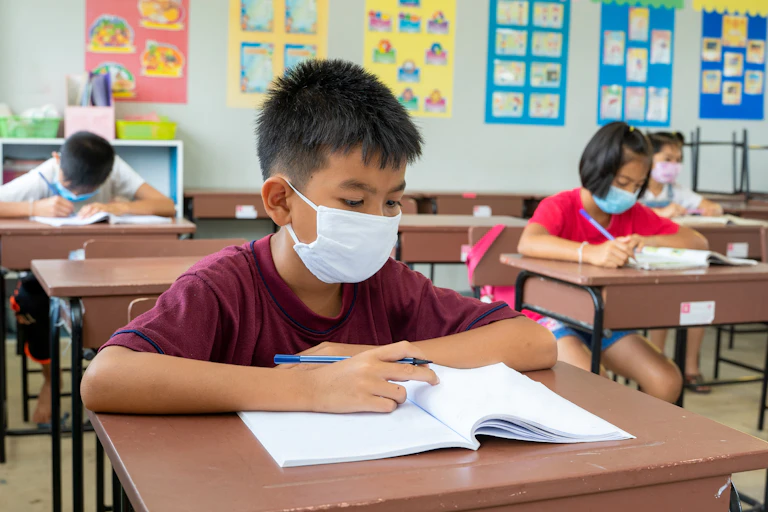 Boy wearing face mask works in notebook at school desk in classroom.