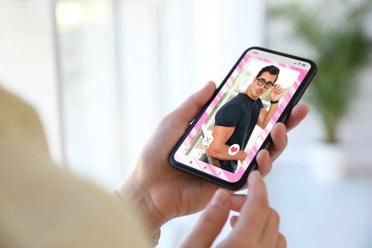 A woman's hand holds a phone showing a photograph of a man wearing glasses and a black muscle tee