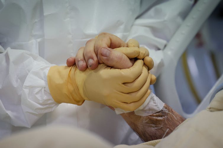An ICU worker in yellow gloves holds a patient's hand.