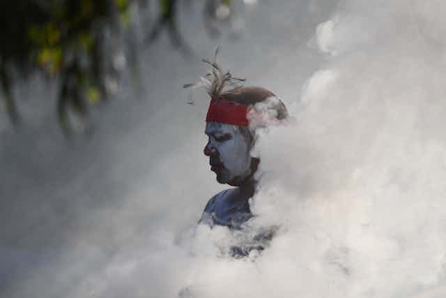 An Aboriginal person with white ochre on their face is submerged in smoke during ceremony.