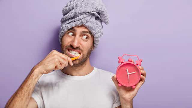 Man with towel on his head brushes his teeth while looking at a clock.